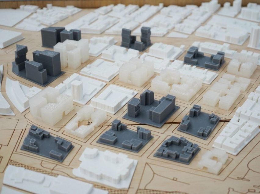 A photograph of an architectural model showing multiple different buildings in tones of grey and white, placed on a brown wooden surface.