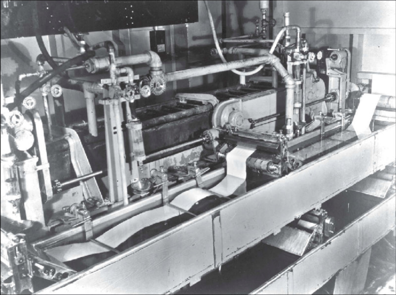 A vintage black-and-white photo of an industrial printing press. The machine is equipped with numerous pipes, valves, and rollers, feeding a continuous roll of paper. The setting appears to be a factory environment.