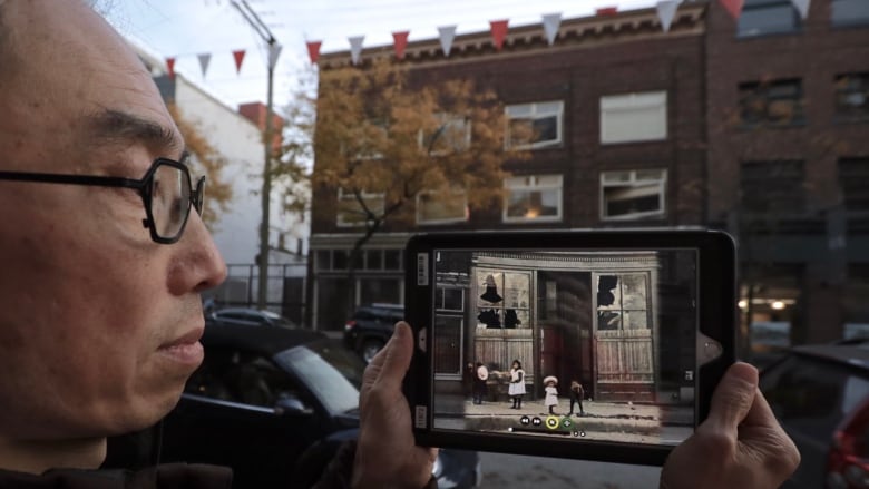 An Asian man holds an iPad in front of a Vancouver street. On the iPad, there's a photo of the street years earlier with destroyed windows and children in front.