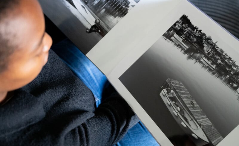 A person wearing a black sweater looks at a photo album, focusing on a black and white image of a docked boat on calm water with buildings in the background.