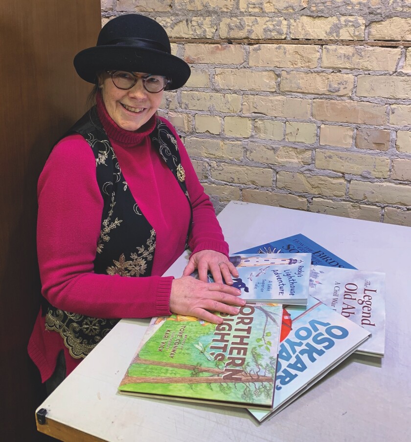 Light-skinned woman sits at table with stack of picture books, smiling. She wears plum-read shirt, flowered vest and black hat.