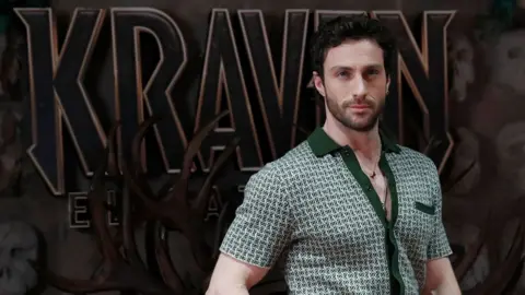 EPA Aaron Taylor-Johnson posing in front of a large Kraven logo at a premiere