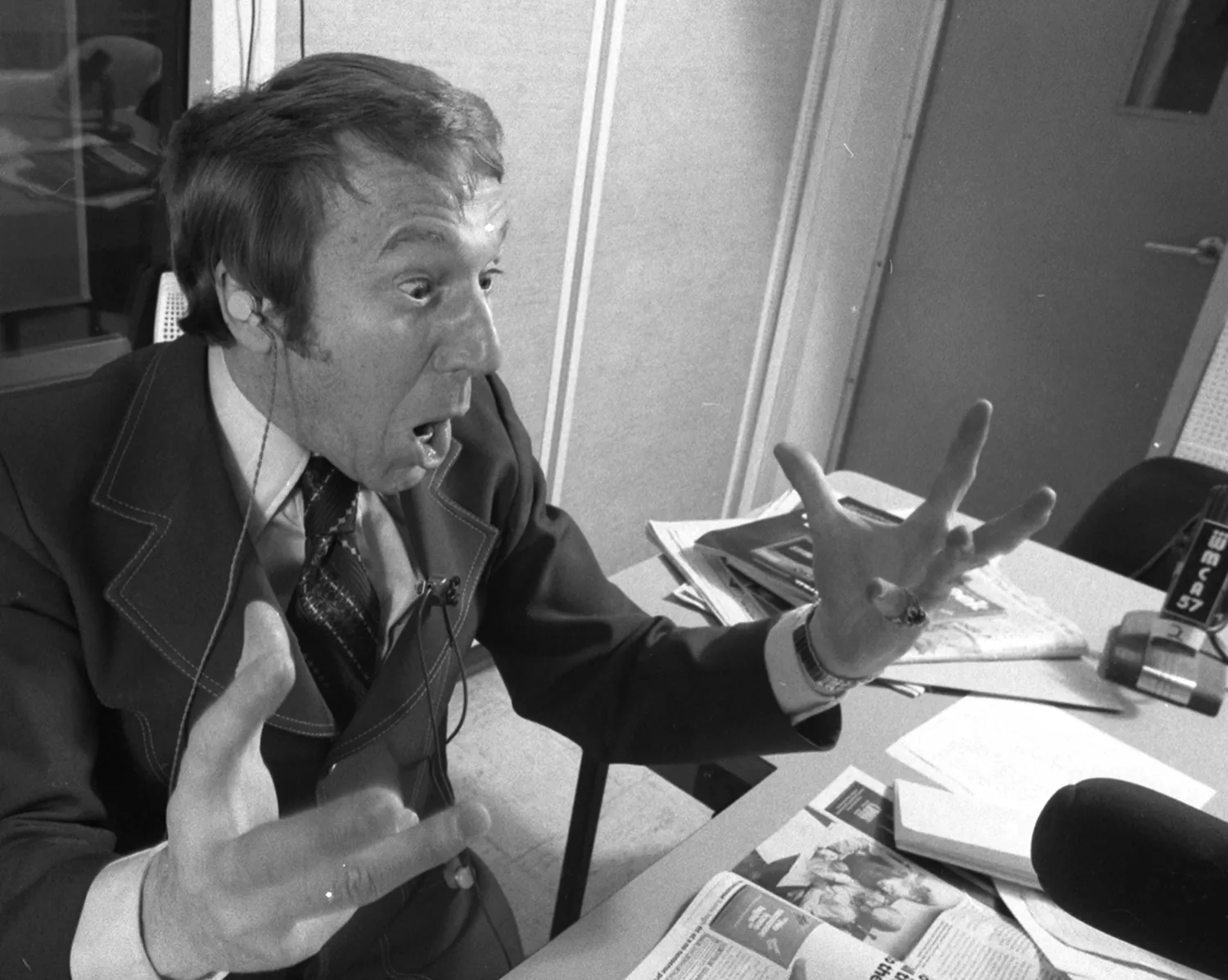 In a historic black-and-white photograph, radio host Bob Grant sits at a desk and appears to be yelling as he gestures with animated hands. His eyes and mouth are wide open.