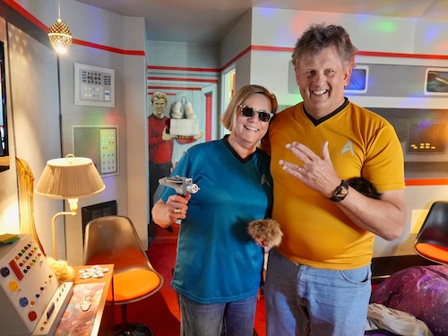 man and woman, both in Star Trek Starfleet shirts, smile in a motel room decorated like the Starship Enterprise