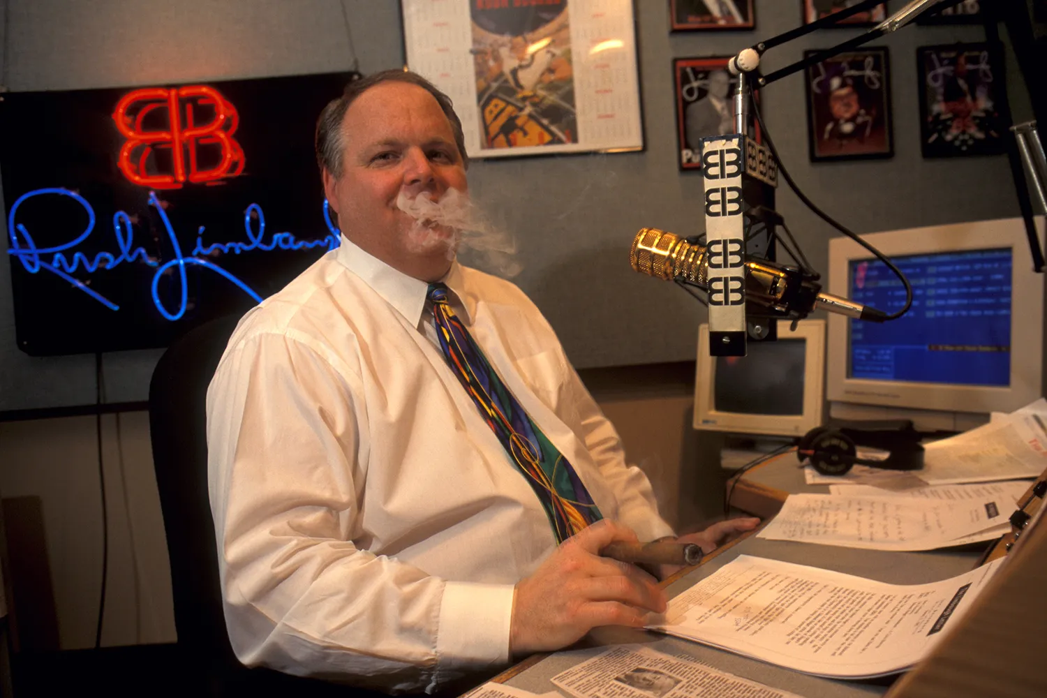 Rush Limbaugh wears a white button-up shirt with a tie as he smokes a cigar in a radio studio, smoke wafting about in a cloud around his head. A microphone, notes, and blocky desktop computer are visible on the desk in front of him.