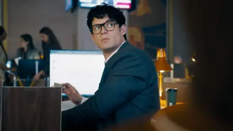 DC Studios/WB David Corenswet as Clark Kent sitting at a desk in the Daily Planet office in a still from the trailer