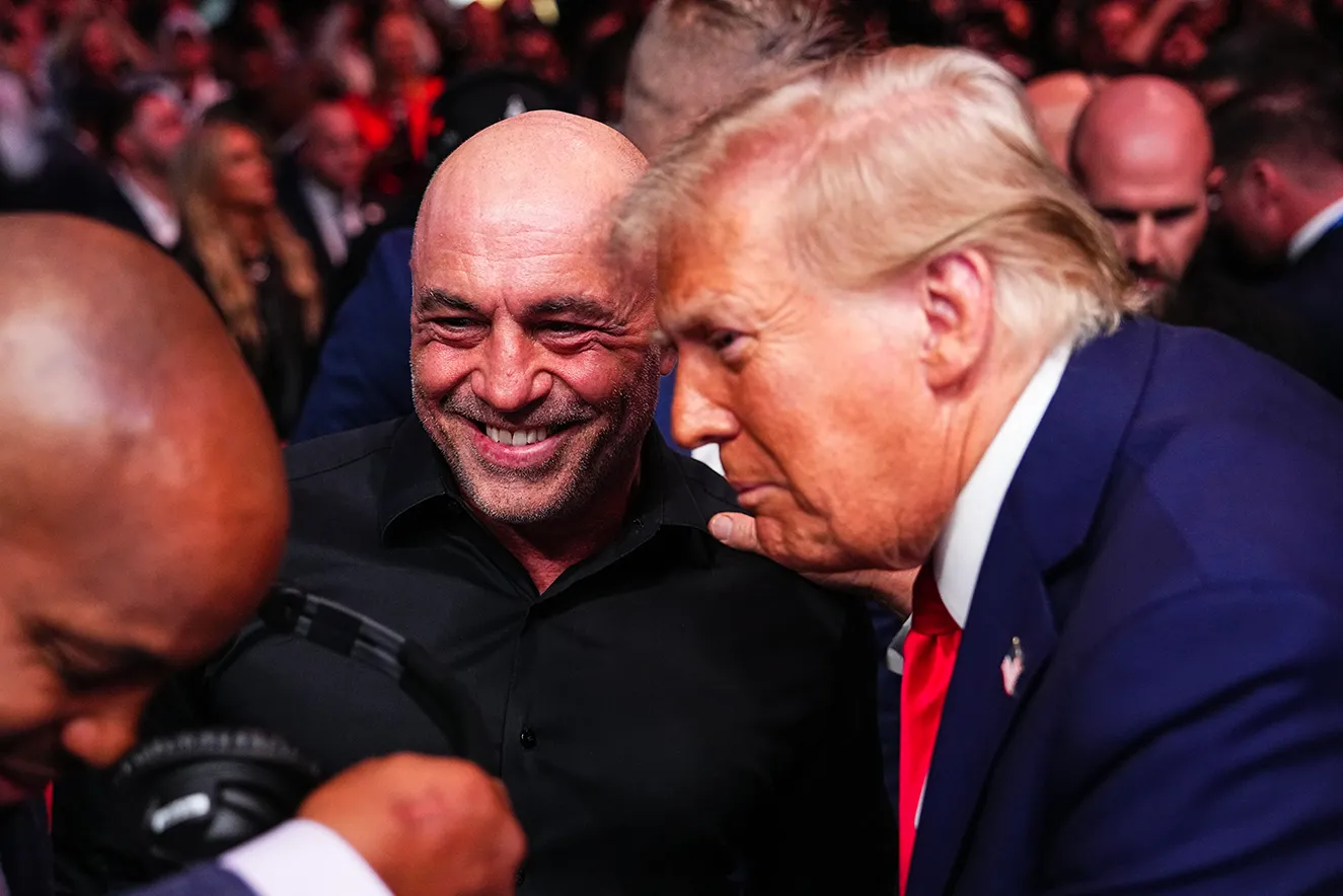 Donald Trump leans in close to talk to Joe Rogan, a crowd of people visible behind them. Trump wears a navy blue suit and red tie, while Rogan wears a black button-up shirt.