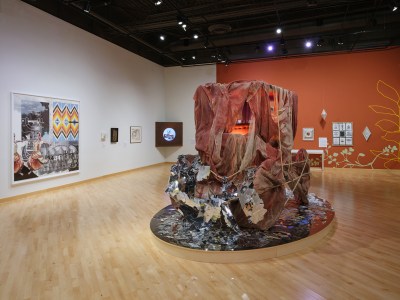 Museum installation view with a large sculpture of a jeep-like car that is covered in fabric and other materials, as wellas several artworks hung on walls in the background.