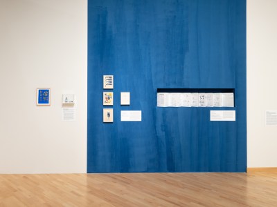 A blue-painted section of a wall contains drawings and a cut-out with a folded brochure. To the left is a small vial of blue pigment. 