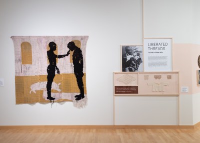 Museum installation view showing a textile artwork with two figures and a wall-hung vitrine with textile samples. 