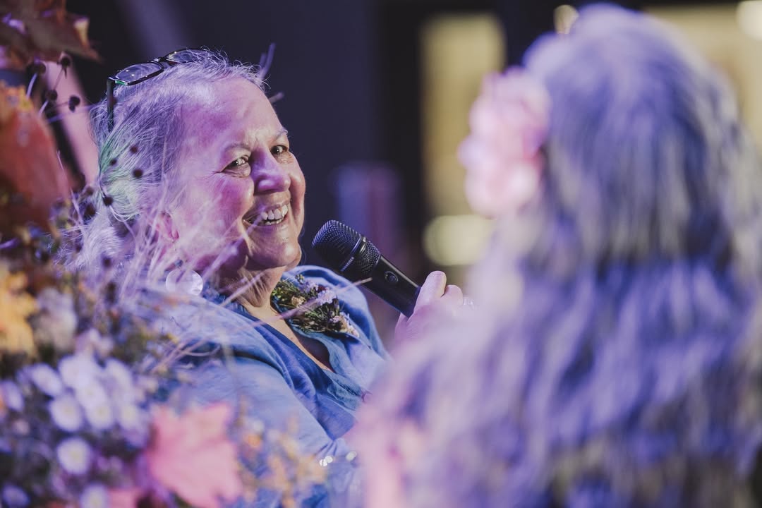 Robin Wall Kimmerer smiling and speaking into a microphone, framed by flowers.