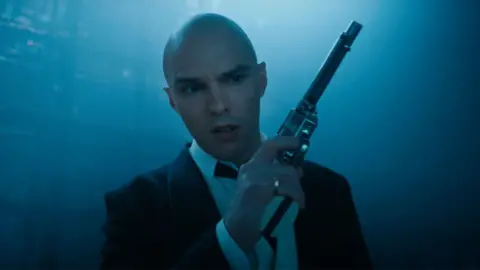 DC Studios/WB A bald Nicholas Hoult in a black suit holding a gun in a still from the trailer