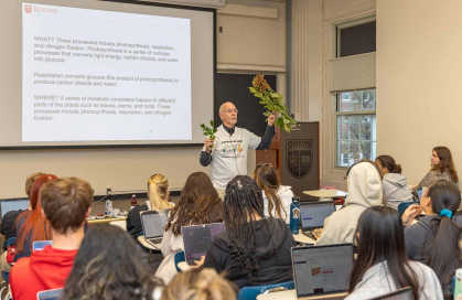 Professor stands next to slide speaking to class