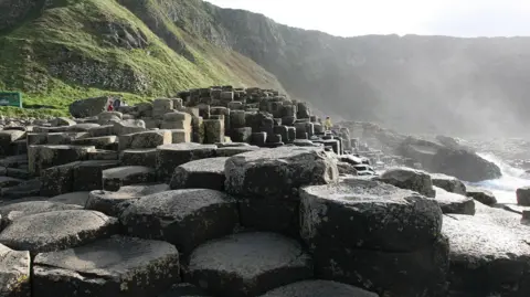 PA Giants Causeway - several dozen dark, circular rocks. Behind the rocks, there is a large cliff-face with plenty of grass. On the right, the sea is meeting the rocks. It is a sunny day.