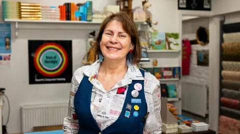 Diane Park A woman with short brown hair wearing a white shirt with printed text on it and a navy blue waistcoat with badges on. She is standing in a bookshop. 