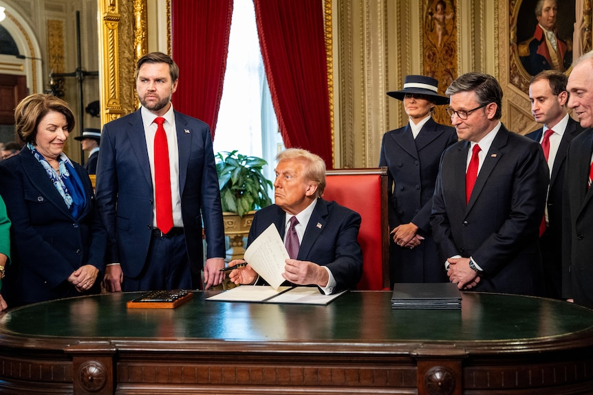 Donald Trump sits at the Resolute Desk with JD Vance on his left and Melania on his right.