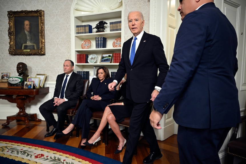 An elderly man in a suit strides into a round room as a middle-aged man and woman sit on chairs beside the door.