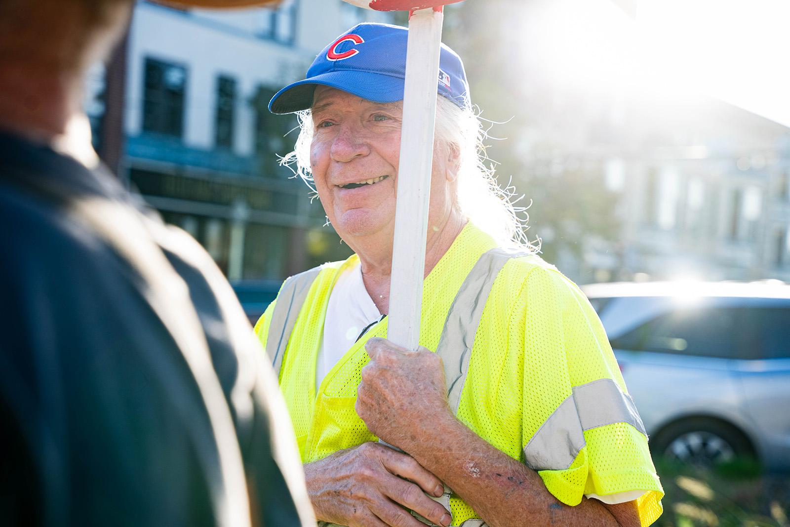 A man in a cubs hat and a yellow safety vest smiles while holding up a sign.