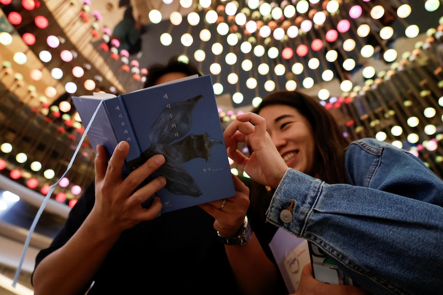 A woman smiles and turns a page of a book held by her companion with blurry orbs of light on the ceiling behind them.