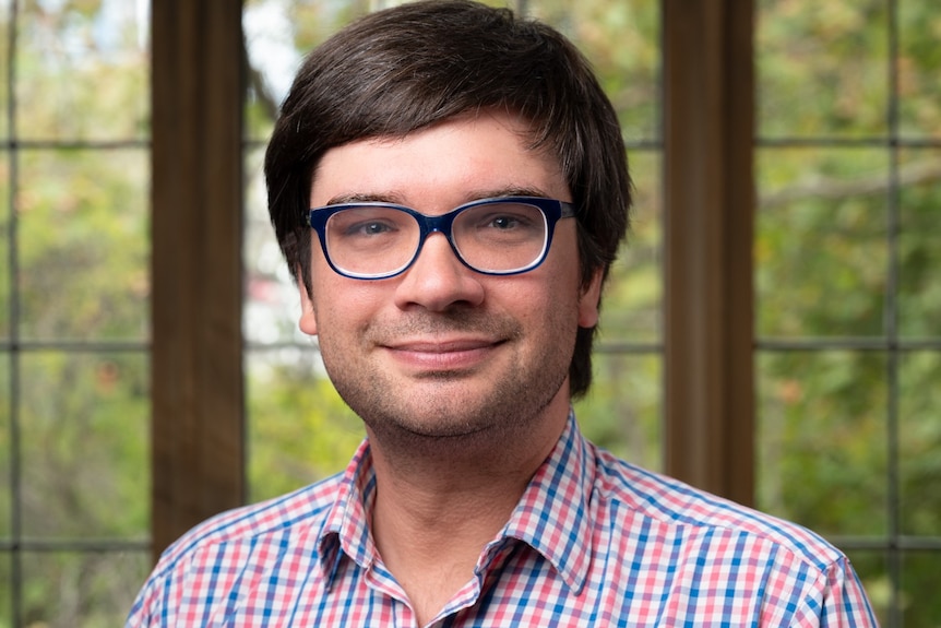 An academic man with glasses and short brown hair smiling with windows and trees in the background.