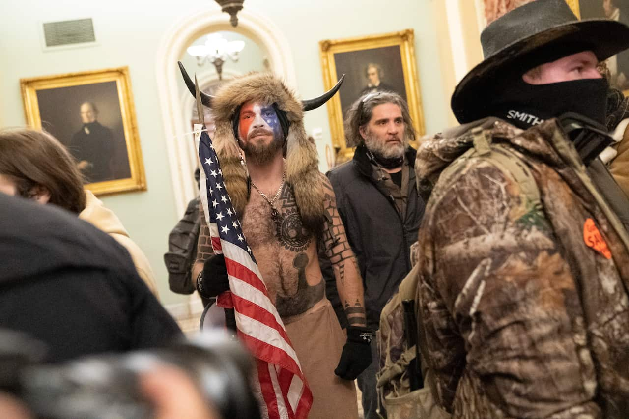 A man wearing a fur headgear with horns on it. His face is painted red, white and blue and he is holding a US flag. His chest is bare