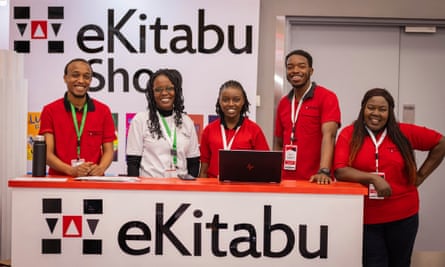 Two men in red T-shirts, two women in red T-shirts and a woman in a white T-shirt, all wearing lanyards, stand behind a desk that reads ‘eKitabu’