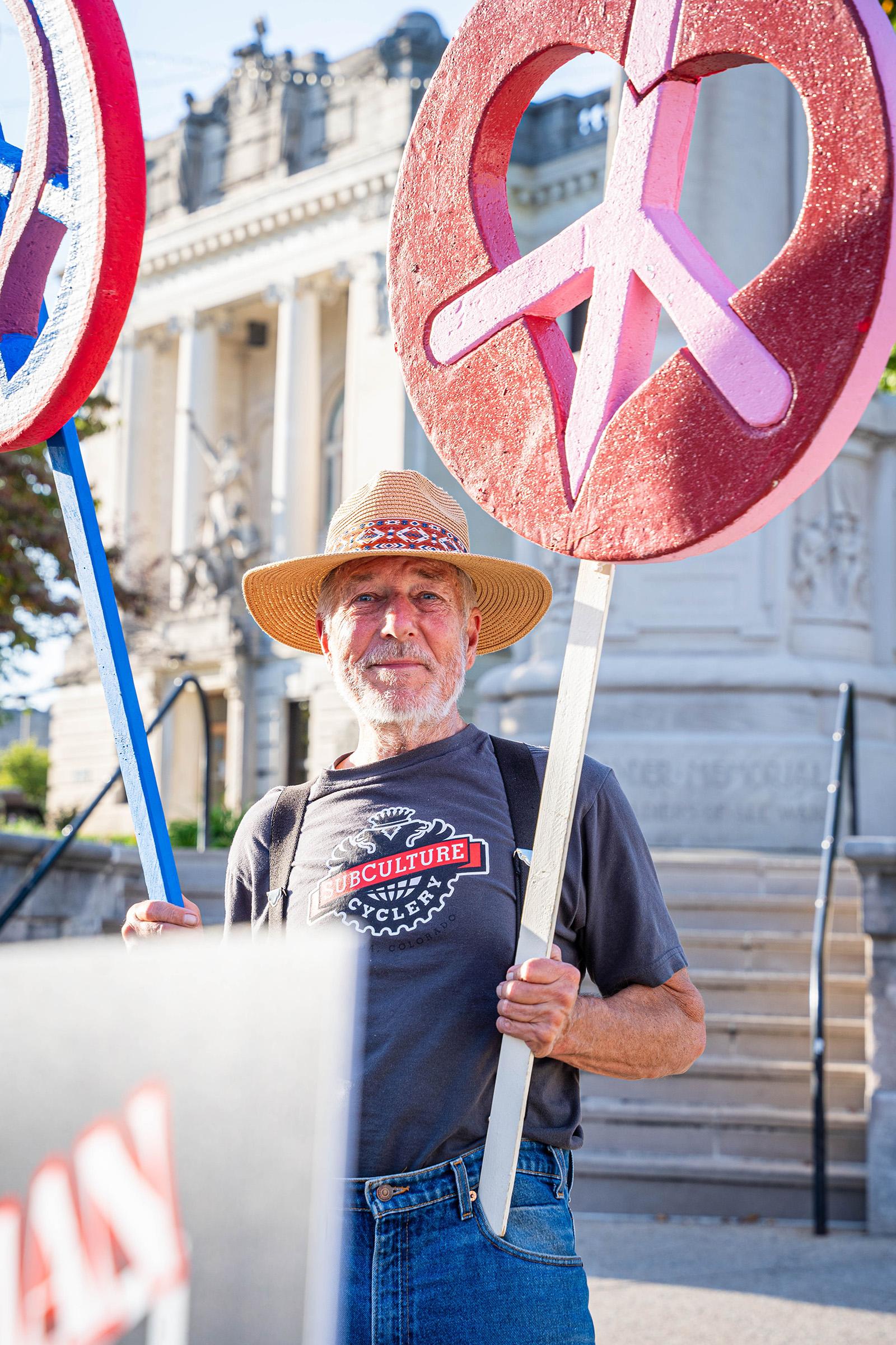 A man in a straw hat smiles at the camera while holding two large peace signs out of frame.