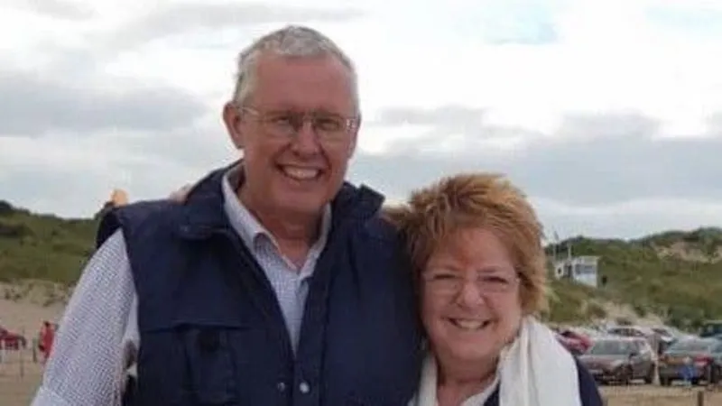 Handout A couple with their arms around each other in front of sand dunes with cars in the background. The sky is cloudy and the couple are smiling. The man is wearing a navy gilet over a white shirt with grey short hair and glasses and the woman has ginger windswept spiky long hair with glasses and she is wearing a white scarf. 