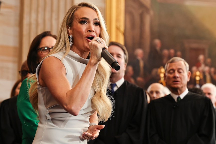 Carrie Underwood sings into a microphone, wearing a silver dress, infront of a crowd of people.