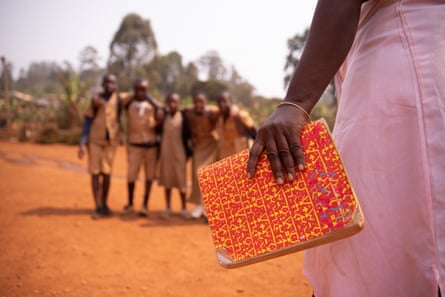 A hand holds an orange book, with children in uniform in the distance out of focus.