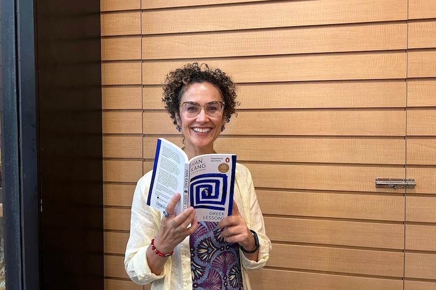 A woman with short curly hair and glasses reading Han Kang's Greek Lessons.