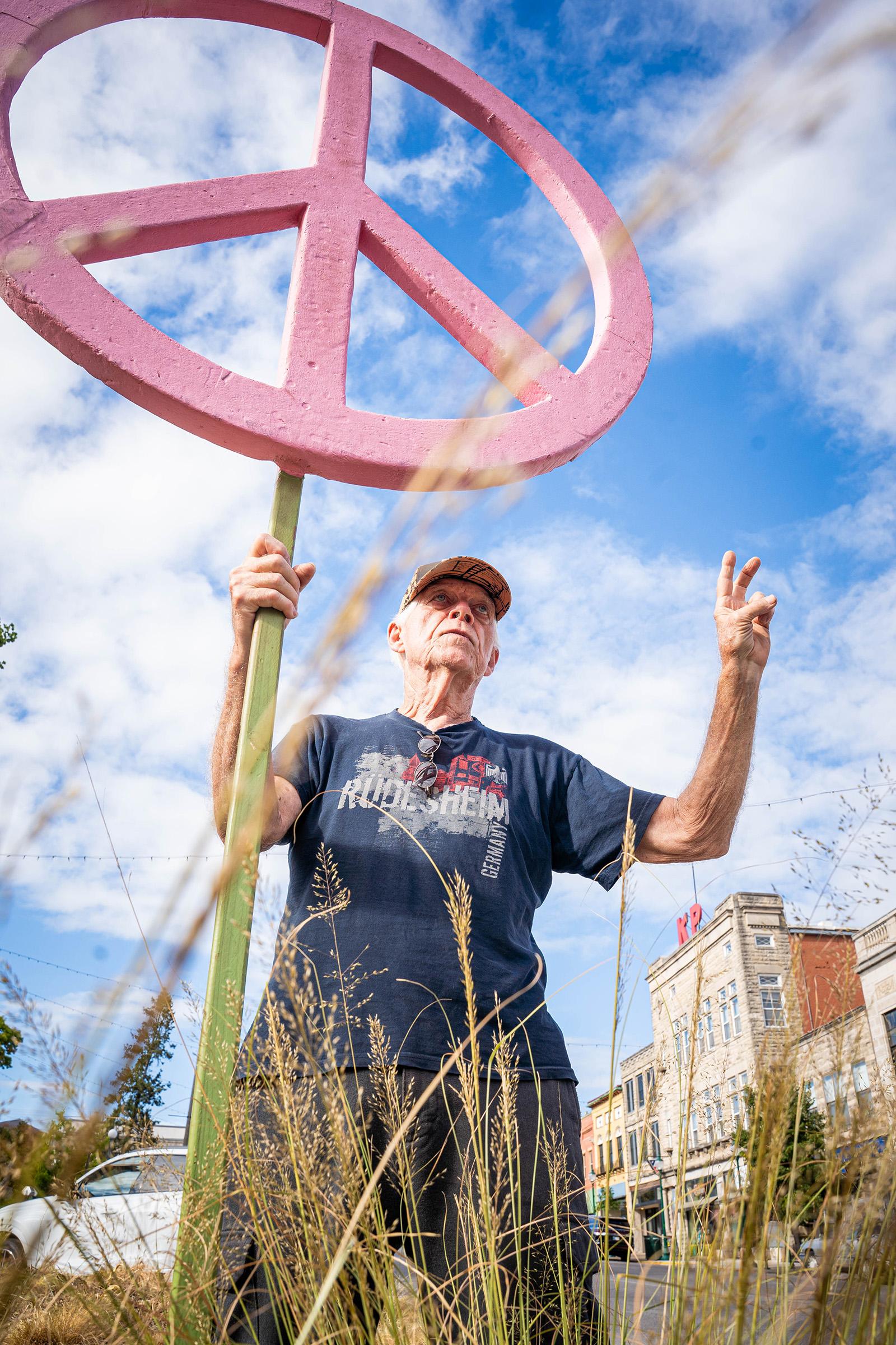 A man holds a big pink peace sign in his right hand while making a peace sign with two fingers with his left hand.