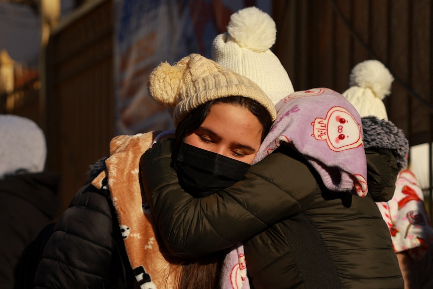 Two women wearing beanies embrace while waiting in line.