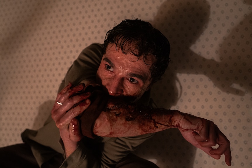 An intense-looking man sits against a wall with his scratched and bleeding arm in his mouth. 