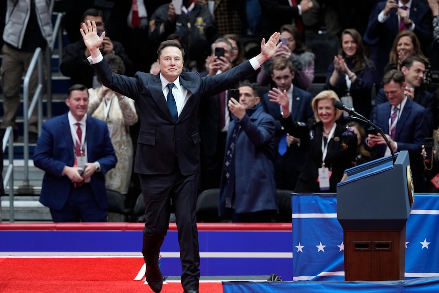 Elon Musk arrives on stage with arms raised in a victory pose to speak at Trump's Inauguration.