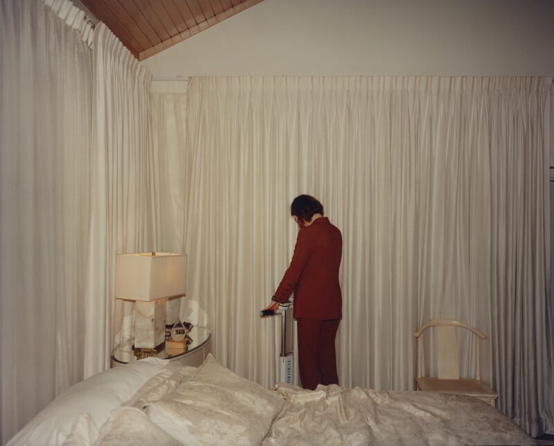 A person in a red suit stands in a room with beige curtains and wooden ceiling. They are facing a white air purifier and holding a handle. A bed with white linens and a lamp with a beige shade are on the left. A wooden chair is nearby.