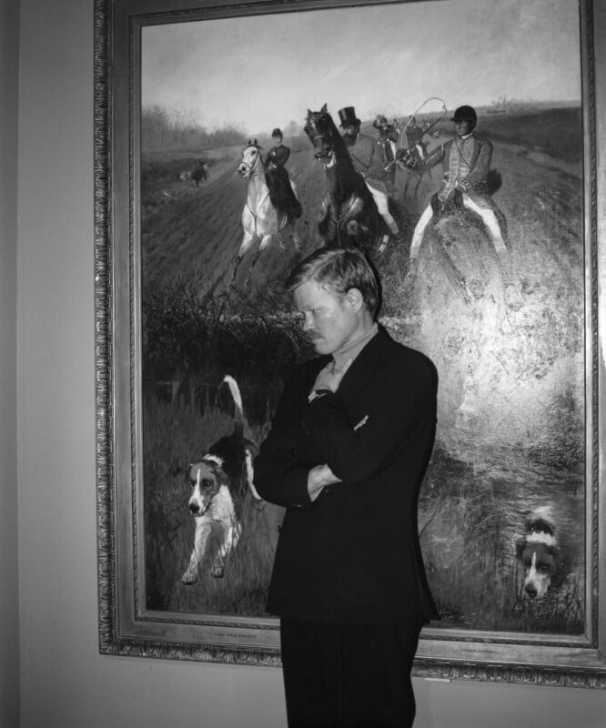 A man in a suit stands pensively in front of a large painting depicting a hunting scene with horses, riders, and dogs in pursuit. The artwork features a dramatic and dynamic outdoor setting.