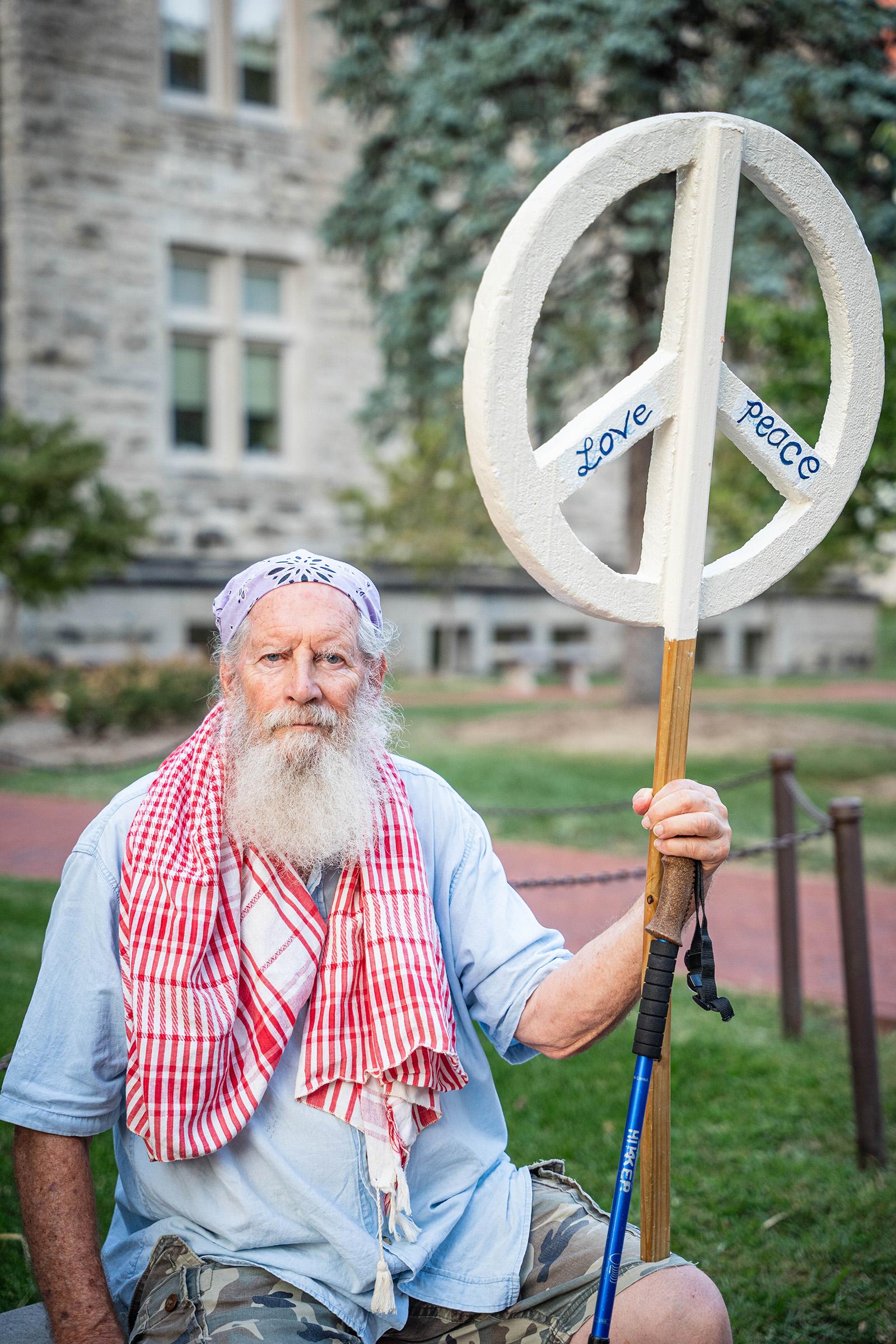 A bearded man holds a big white peace sign with the words 'Love' and 'Peace' on it.