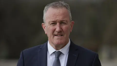 PA Media A man with grey hair smiles in the direction of the camera. He is wearing a dark navy suit with a white shirt and a grey and white tie.