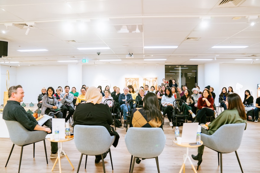 Four people sit with their backs to the camera in chairs facing a crowd of people at a talking panel event.