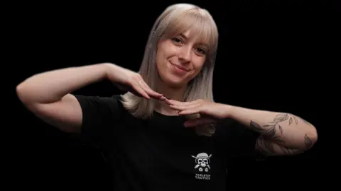 Tabletop Tactics A woman with shoulder length blond hair wearing a black t-shirt poses in front of a black studio background. A white 