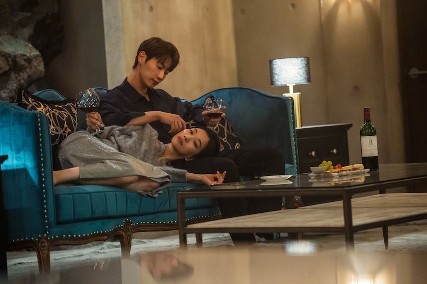 A tilted photo of a Korean woman lying on a couch with a man stroking her hair, wine and cheese on the table in front of them.