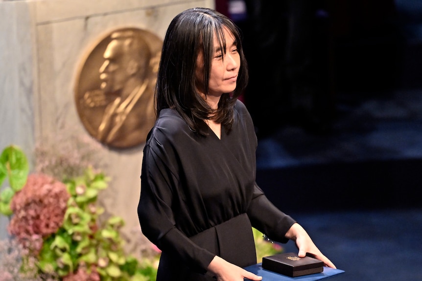 A middle-aged Korean woman holding a book and box in long black dress on stage with Nobel medallian in background.