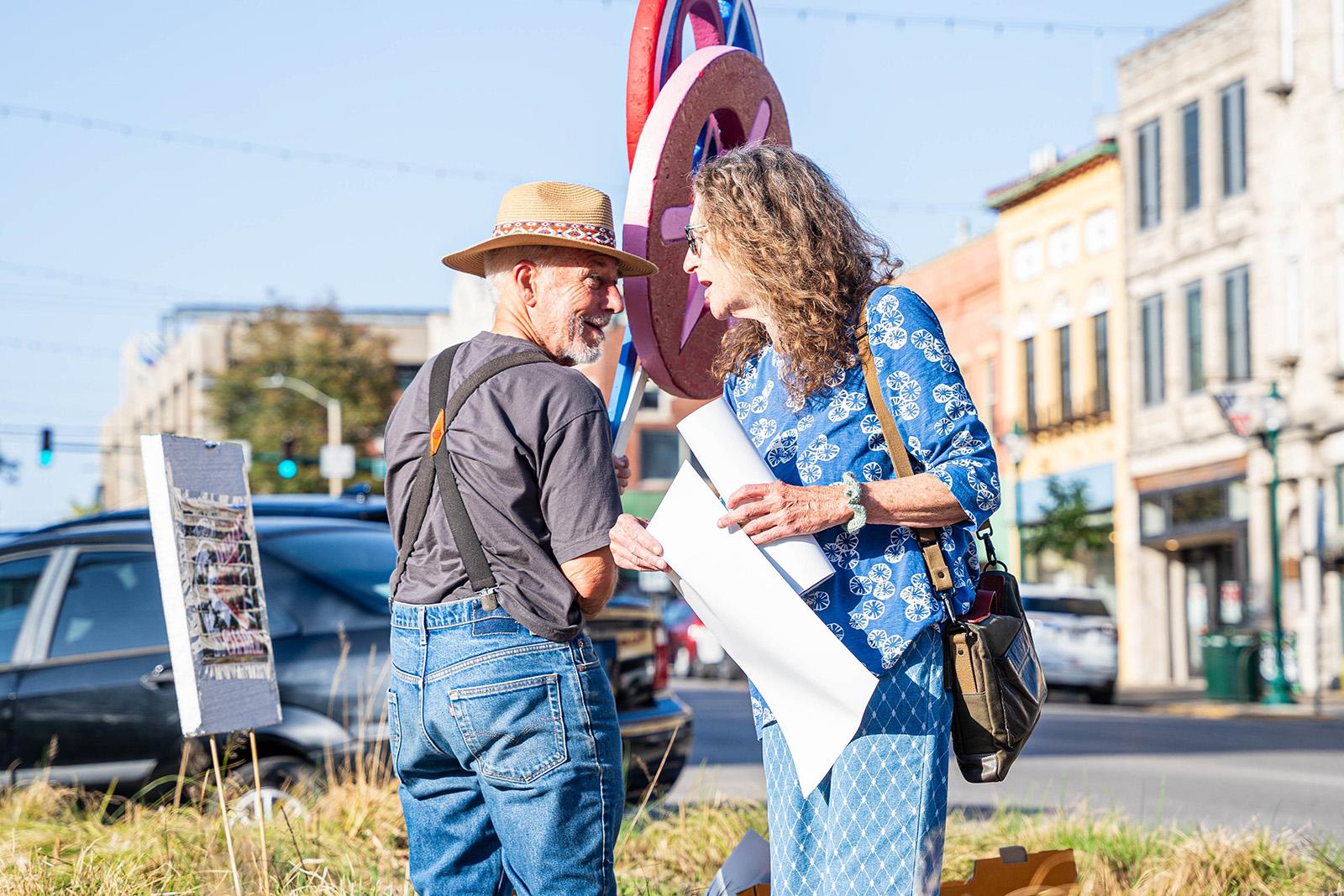 A man in a straw hat speaks to a woman wearing a blue blouse and pants who is walking past him.