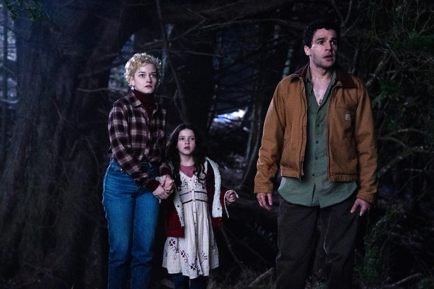 A man, woman and young girl stand outside in a wooded area looking up at something out of frame.