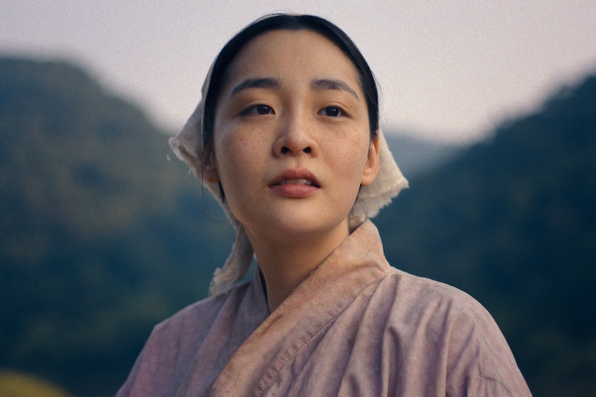 A close up of a Korean woman in soft pink traditional dress and head covering looking wistfully with mountains in background.