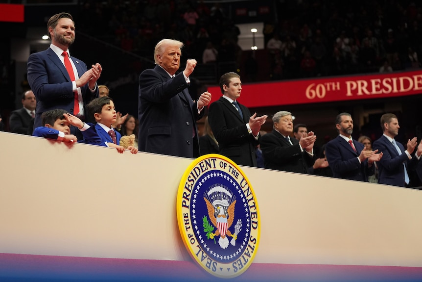 Donald Trump attends an indoor inauguration event, standing above US presidential coat of arms with fist raised.