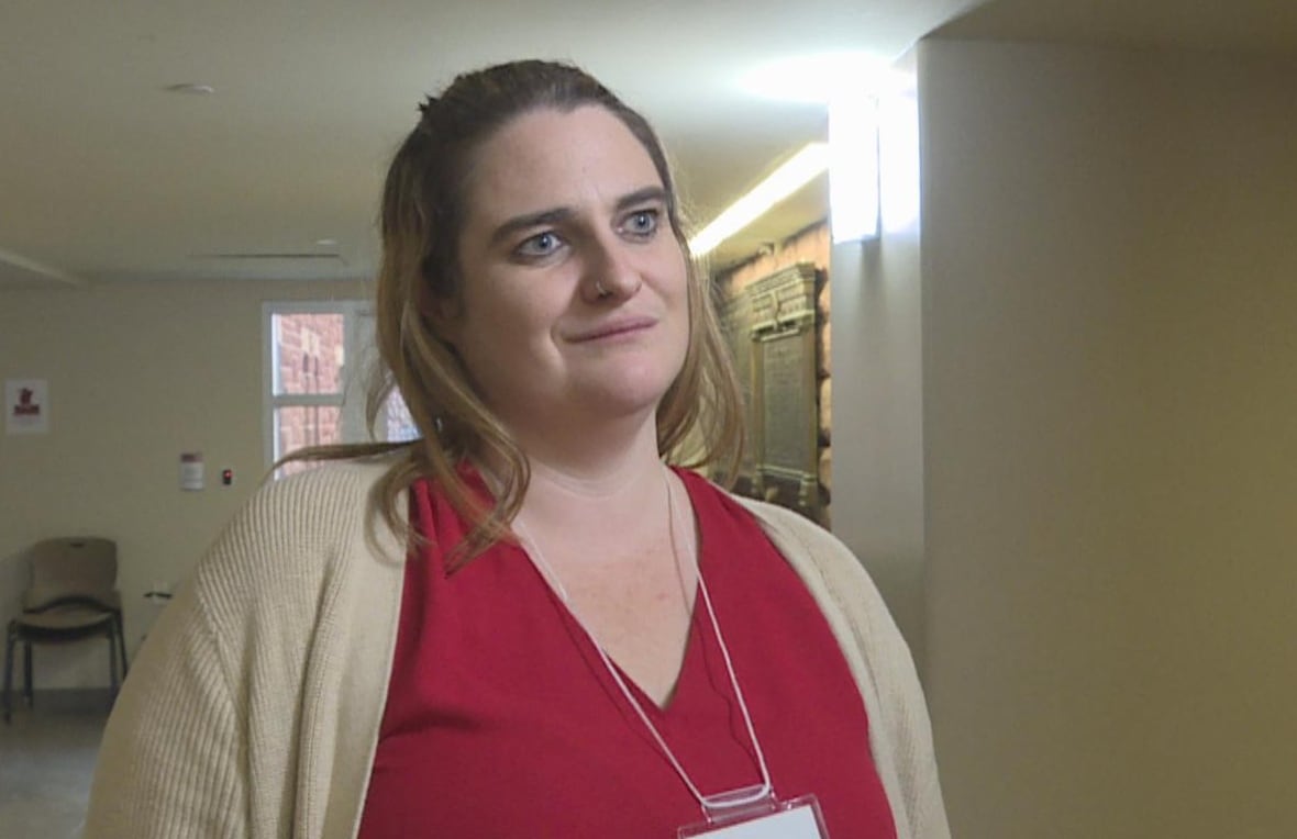 A woman with medium-length, blonde hair stands inside a hallway wearing a red top and beige cardigan.