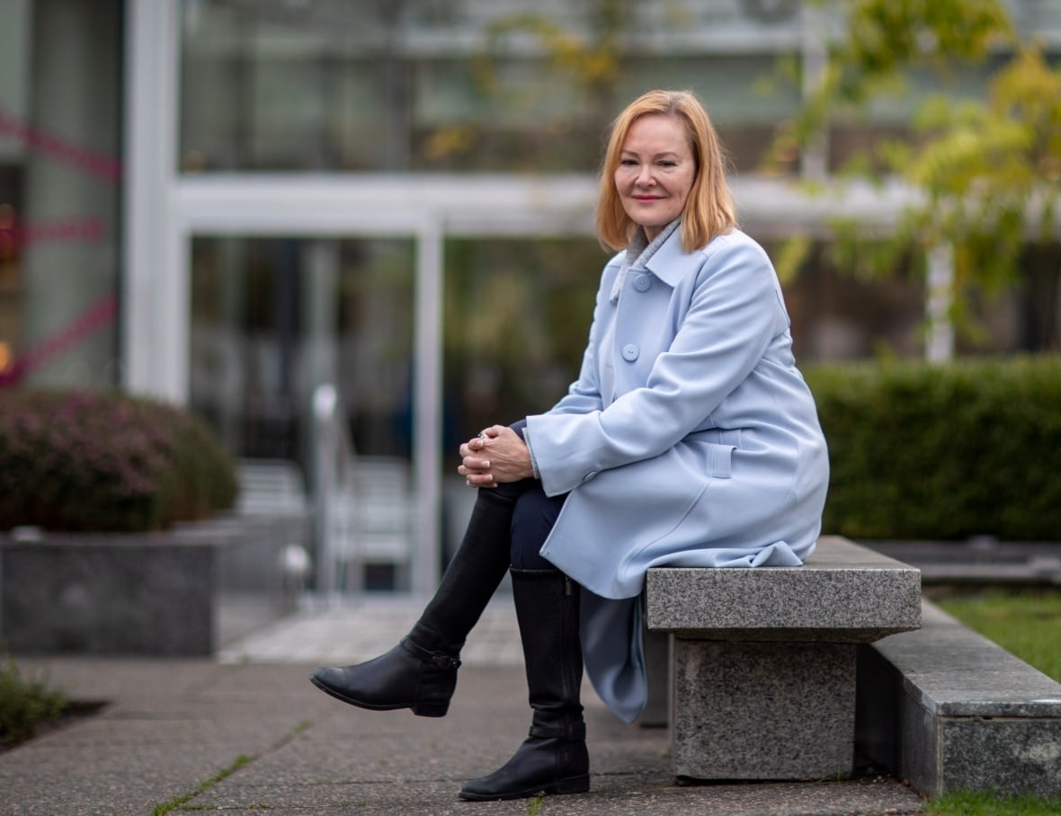 A white woman wearing a blue coat sits on an outdoor bench.