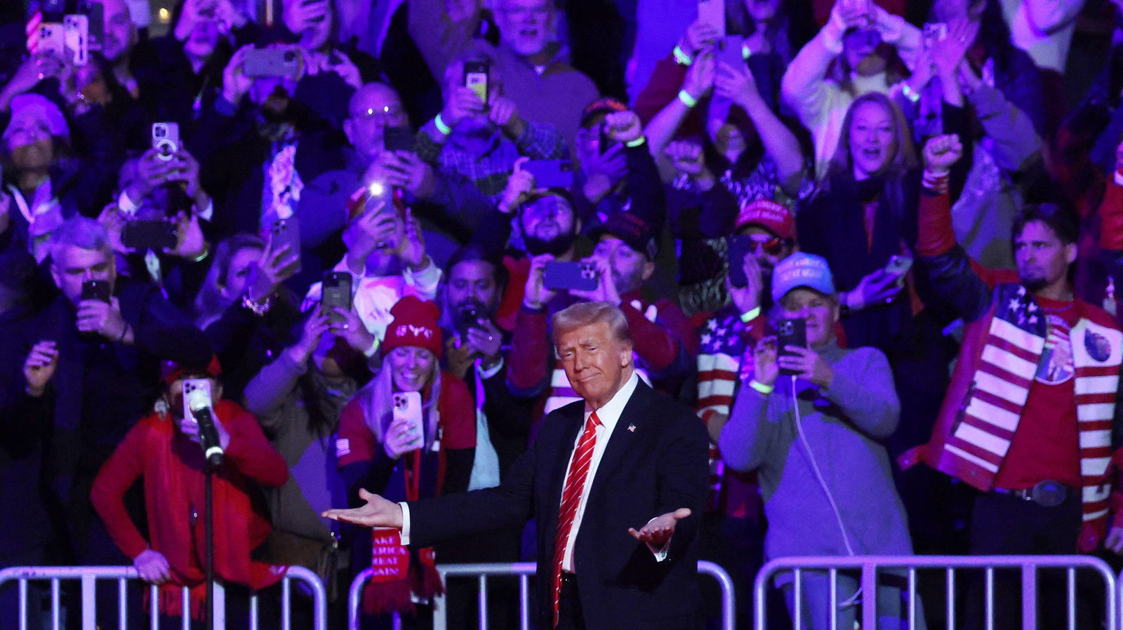 Donald Trump raises both palms in a shrug as a crowd behind him takes photos on their phones 